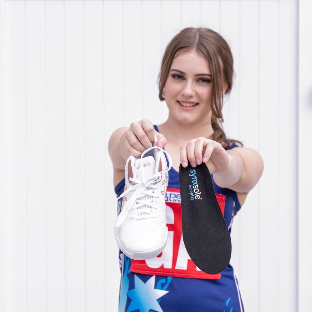 girl wearing netball uniform holding up asics sneakers and synxsole kids insole, severs disease, growing pains